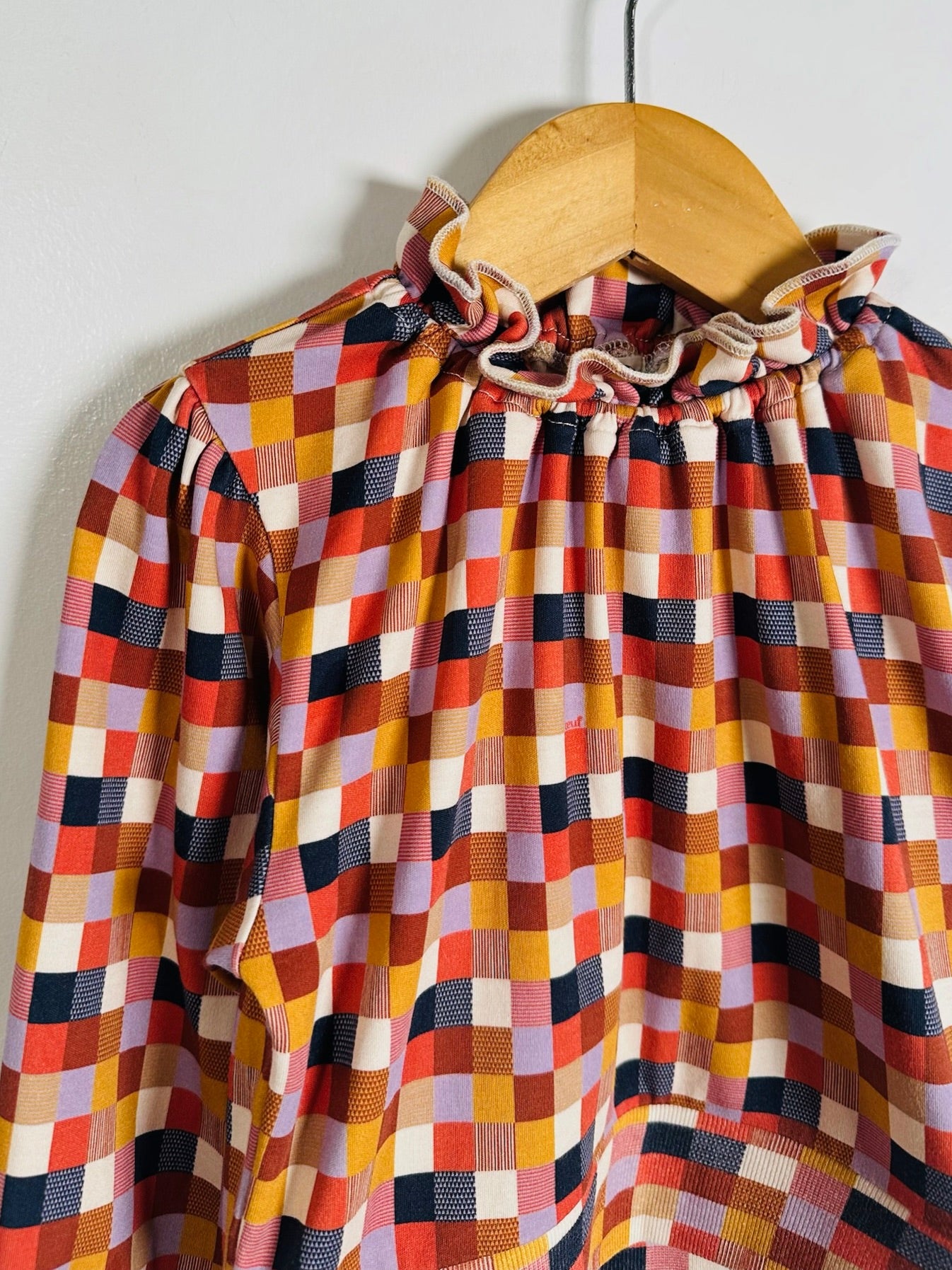 A long-sleeve blouse with a colorful checkered pattern, featuring a ruffled mock neck, hanging on a wooden hanger against a plain light gray wall.