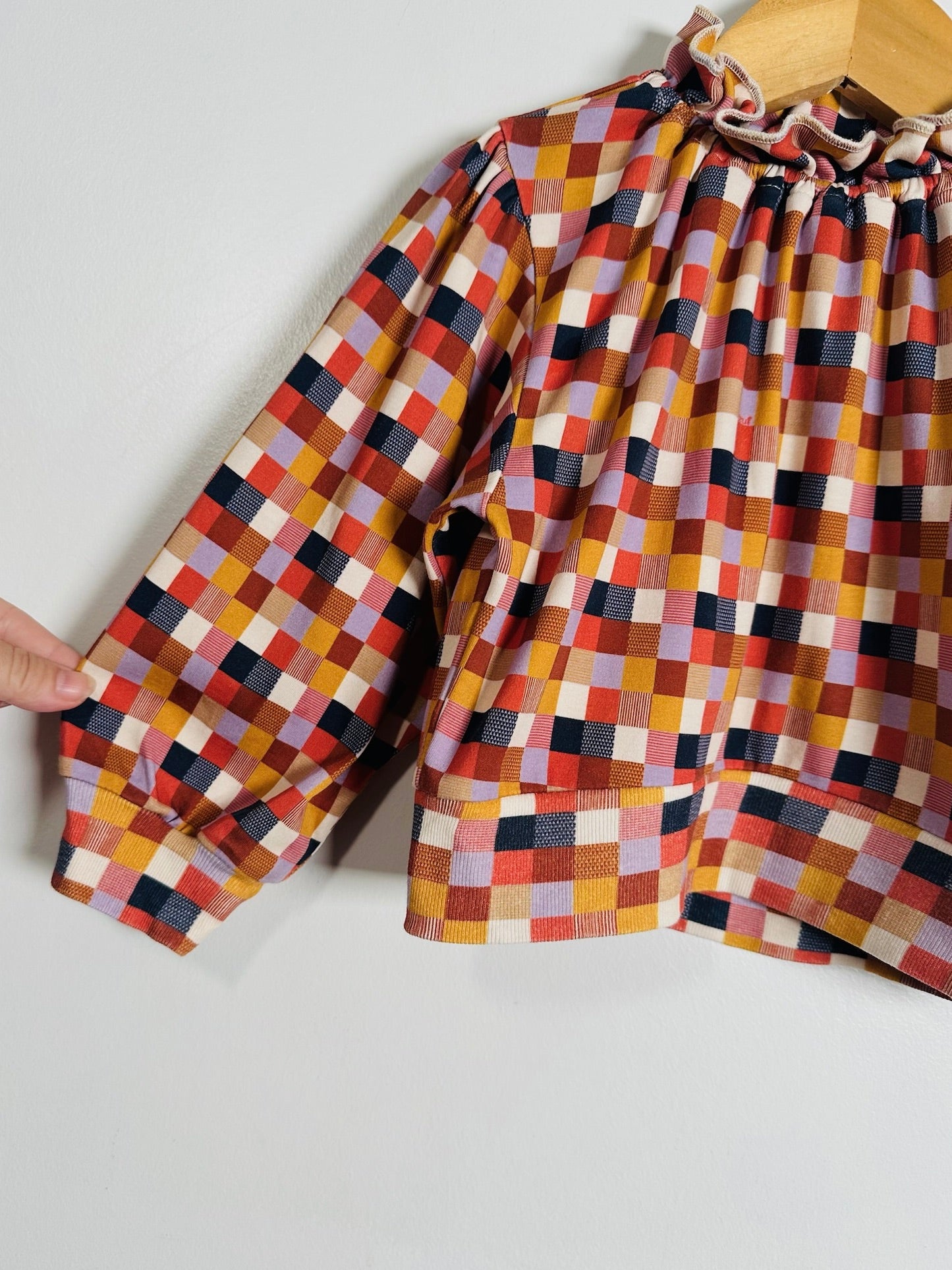 A long-sleeve blouse with a colorful geometric checkered pattern, featuring a ruffled smocked neckline and a pleated gathered waist, shown hanging on a wooden hanger against a plain light gray wall.