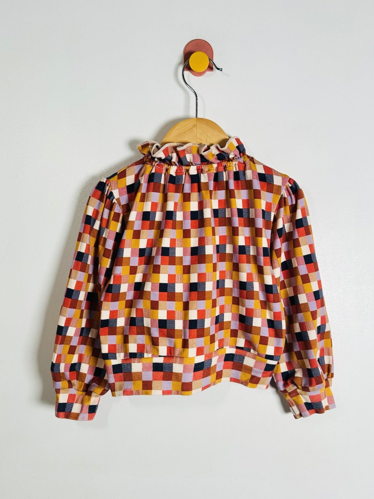 A long-sleeve blouse with a colorful geometric checkered pattern, hanging on a wooden hanger from a wall hook against a plain white wall.