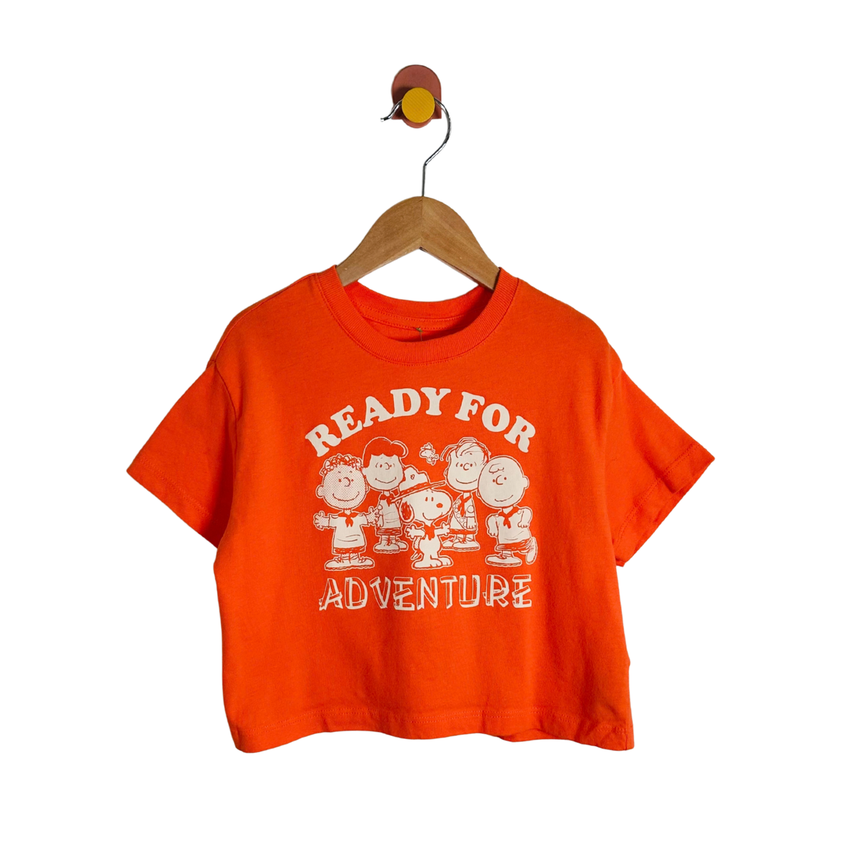 An orange short-sleeve crop top t-shirt featuring a white graphic of the Peanuts characters and the text 'READY FOR ADVENTURE' on the front, hanging on a wooden hanger against a white background.