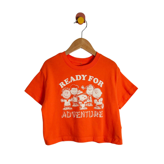 An orange short-sleeve crop top t-shirt featuring a white graphic of the Peanuts characters and the text 'READY FOR ADVENTURE' on the front, hanging on a wooden hanger against a white background.