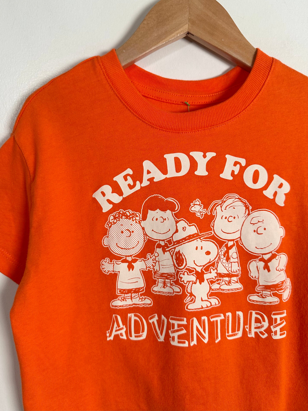 An orange crewneck t-shirt hanging on a wooden hanger against a light wall. The shirt features a white graphic print of the Peanuts characters with the text 'READY FOR ADVENTURE'.
