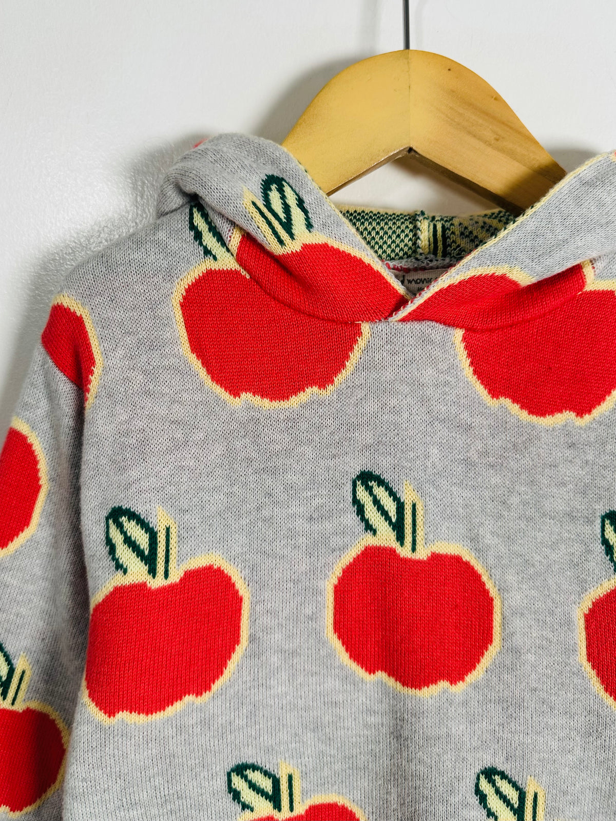 A gray knit hoodie with a repeating red apple pattern outlined in yellow and green leaves, hanging on a wooden hanger against a plain light background.