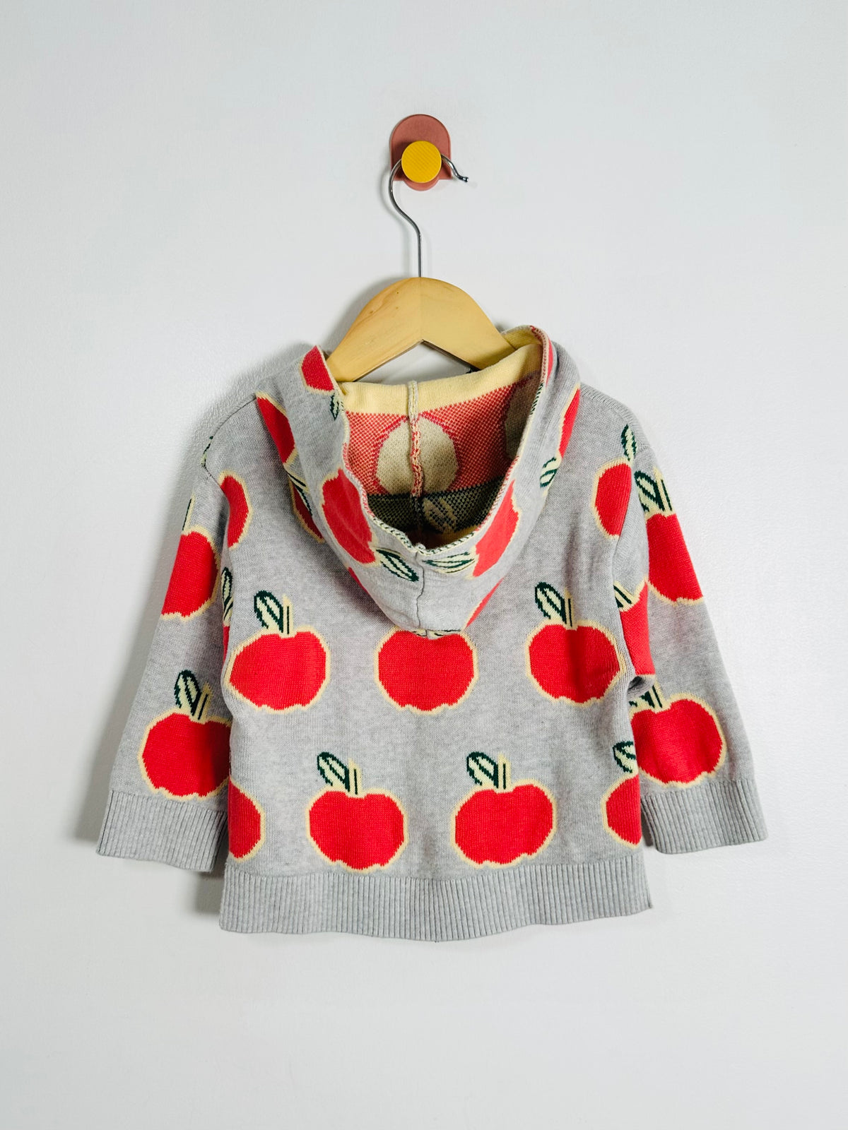A gray knit hooded sweater with a red apple pattern, hanging on a wooden hanger against a plain light gray wall.
