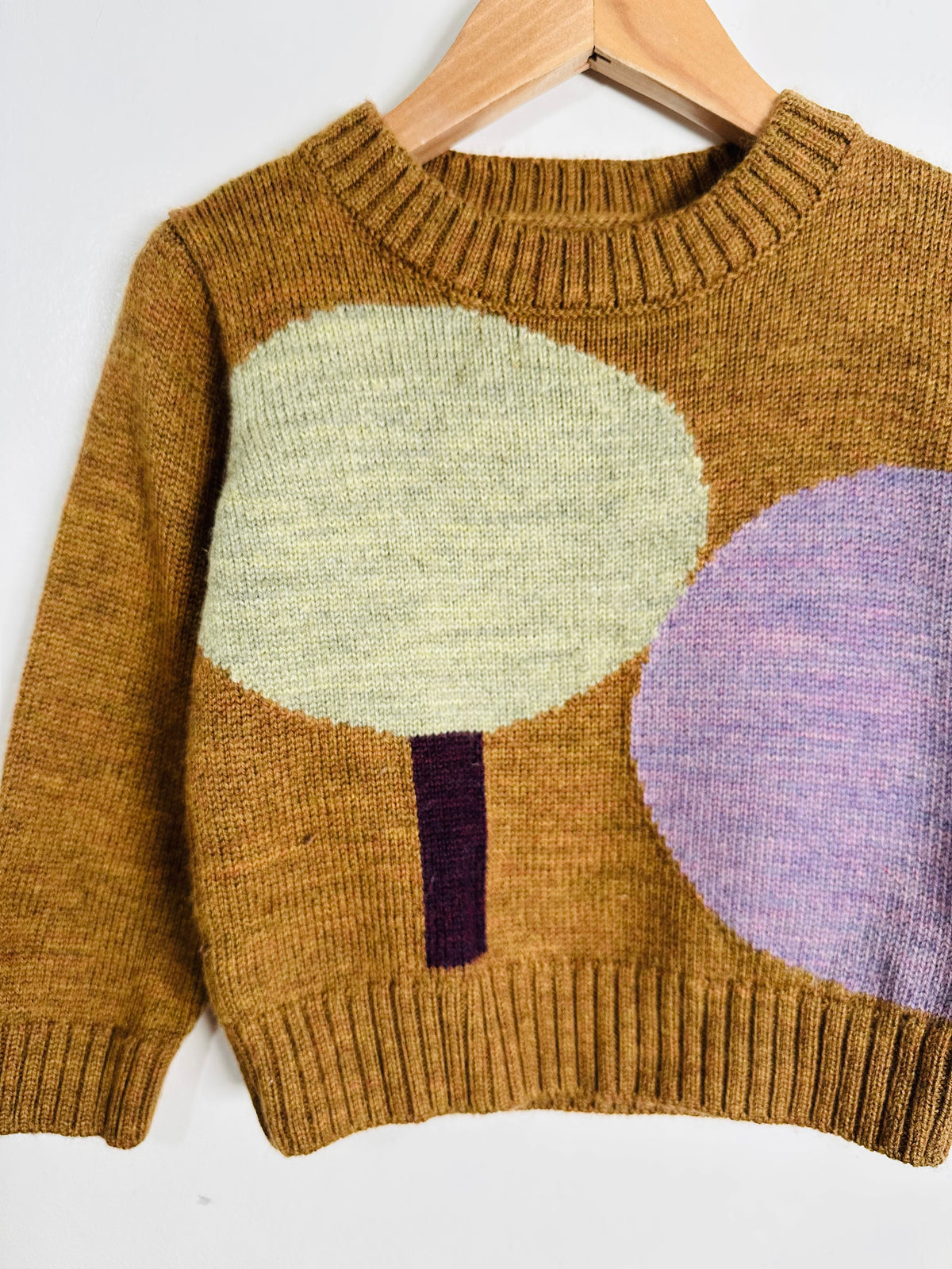 A knit crewneck sweater in a warm camel brown, featuring a large light green tree motif and a purple oval on the front, hanging on a wooden hanger against a white background.