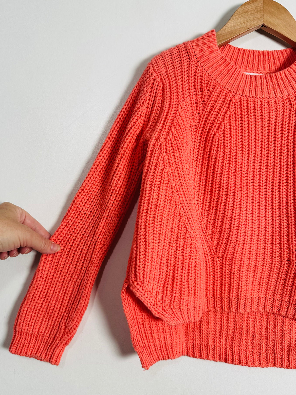 A coral orange chunky knit sweater hanging on a wooden hanger against a light gray wall, with a hand holding the sleeve to show the texture.