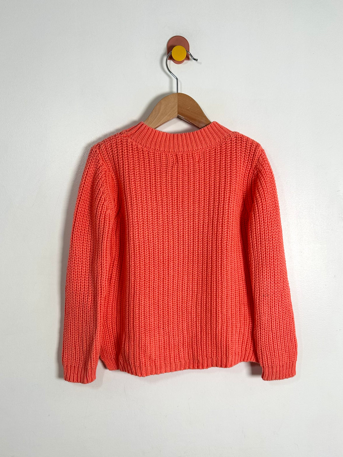 A coral orange ribbed knit sweater hanging on a wooden hanger against a plain white wall.
