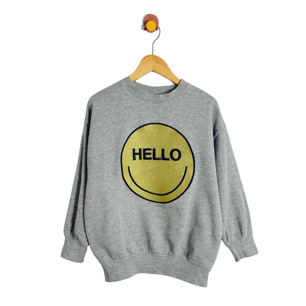 A heather gray crewneck sweatshirt hanging on a wooden hanger against a plain white background. The sweatshirt features a large circular yellow smiley face graphic on the chest with the word 'HELLO' printed inside in black.