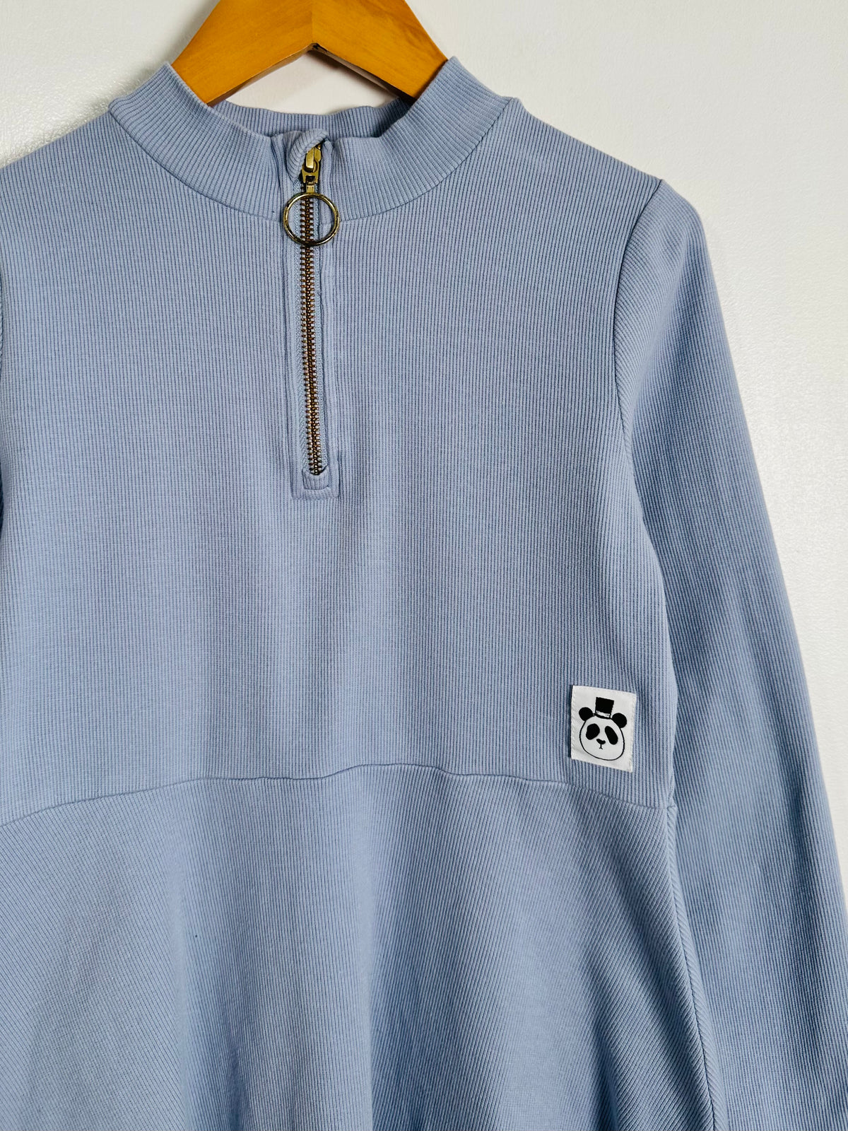 A light blue ribbed long-sleeve quarter-zip pullover hanging on a wooden hanger against a plain off-white wall. The garment features a small white square patch on the left chest with a black panda graphic.