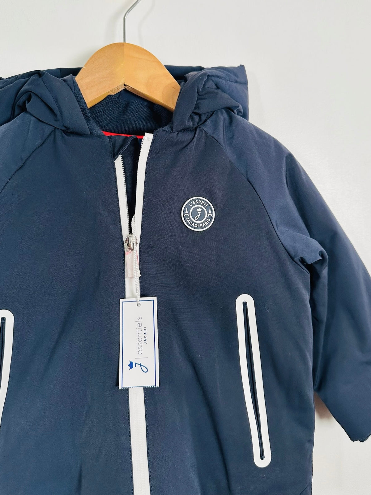 A navy blue hooded jacket hanging on a wooden hanger against a plain light gray wall. The jacket has a white front zipper, white vertical zip pockets, a circular Paris Saint-Germain crest on the left chest, and a hangtag attached to the zipper.