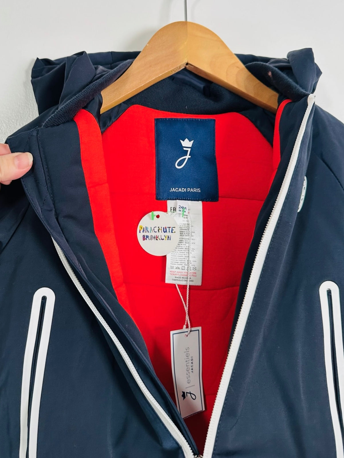 A close-up of a Jacadi Paris hooded jacket hanging on a wooden hanger, showing a navy blue exterior, a bright red interior lining, and white zipper accents. A hand is holding the left shoulder area to display the hood and lining.
