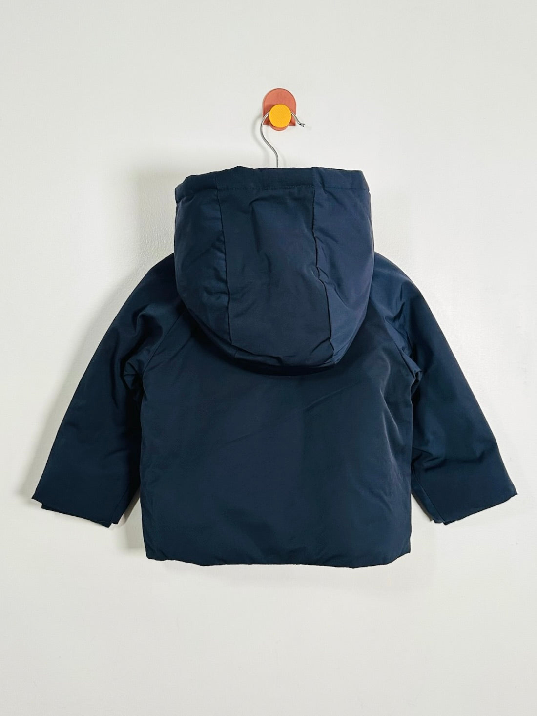 A navy blue hooded puffer jacket shown from the back, hanging on a small orange-and-white hanger against a plain light gray wall.