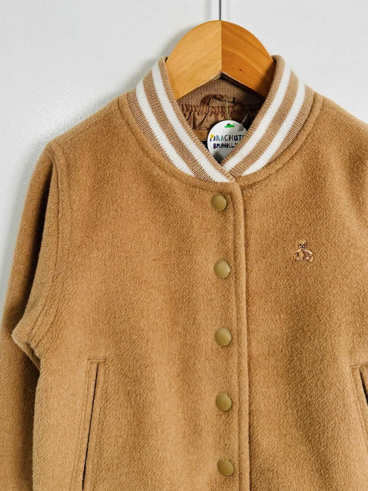 A camel brown wool-blend varsity jacket with cream and brown striped ribbed collar, hanging on a wooden hanger against a white wall.