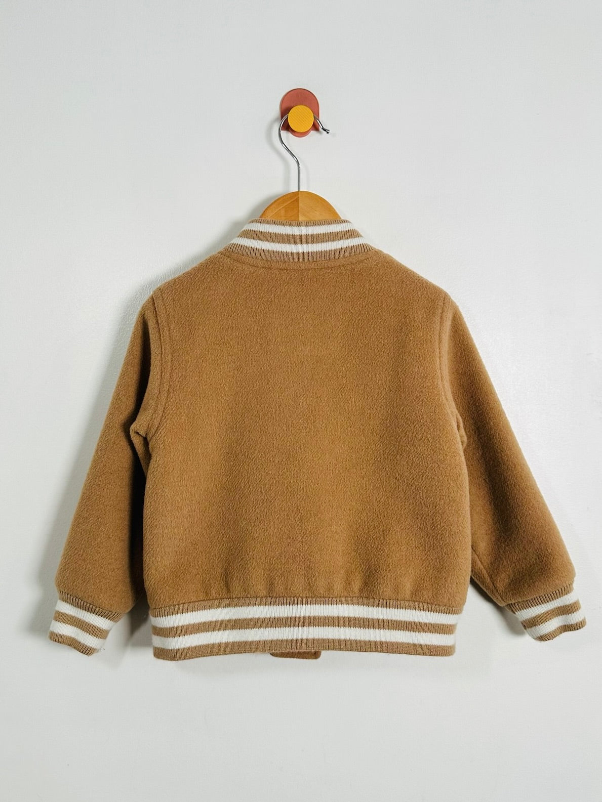 A camel brown knit cardigan sweater with cream and white striped ribbed collar, cuffs, and hem, hanging on a wooden hanger against a light gray wall.
