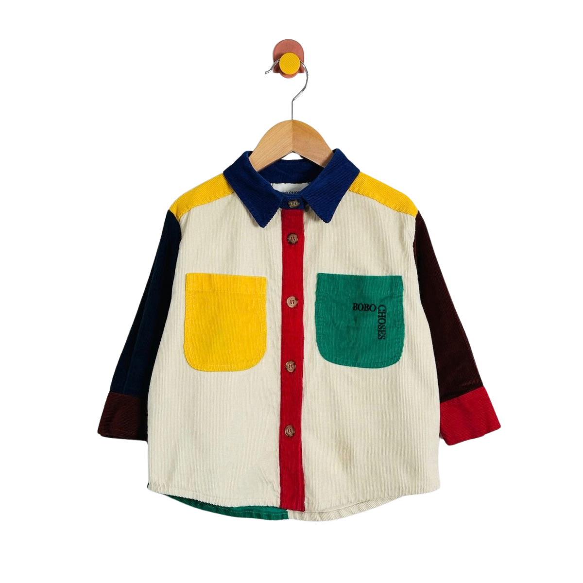 A multicolor patchwork long-sleeve button-up shirt for toddlers, featuring a cream body, yellow shoulder panel, green pocket, red front placket, and navy collar, hanging on a wooden hanger against a white background.