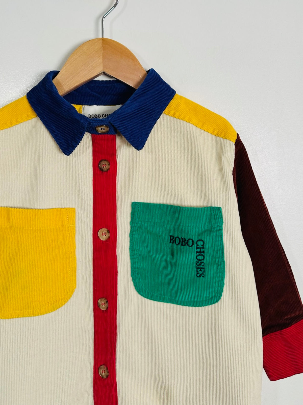 A multicolor corduroy button-up shirt by Bobo Choses hanging on a wooden hanger against a plain white wall. The shirt features a cream-colored body, a blue corduroy collar, a red corduroy placket, a yellow pocket and shoulder panel, and a green pocket on the chest with 'BOBO CHOSES' embroidered vertically.