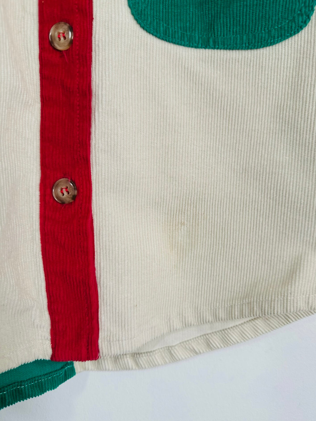 Close-up of a vintage corduroy garment featuring an off-white ribbed body, a red vertical stripe with metal buttons, and green contrast panels at the neckline and hem.