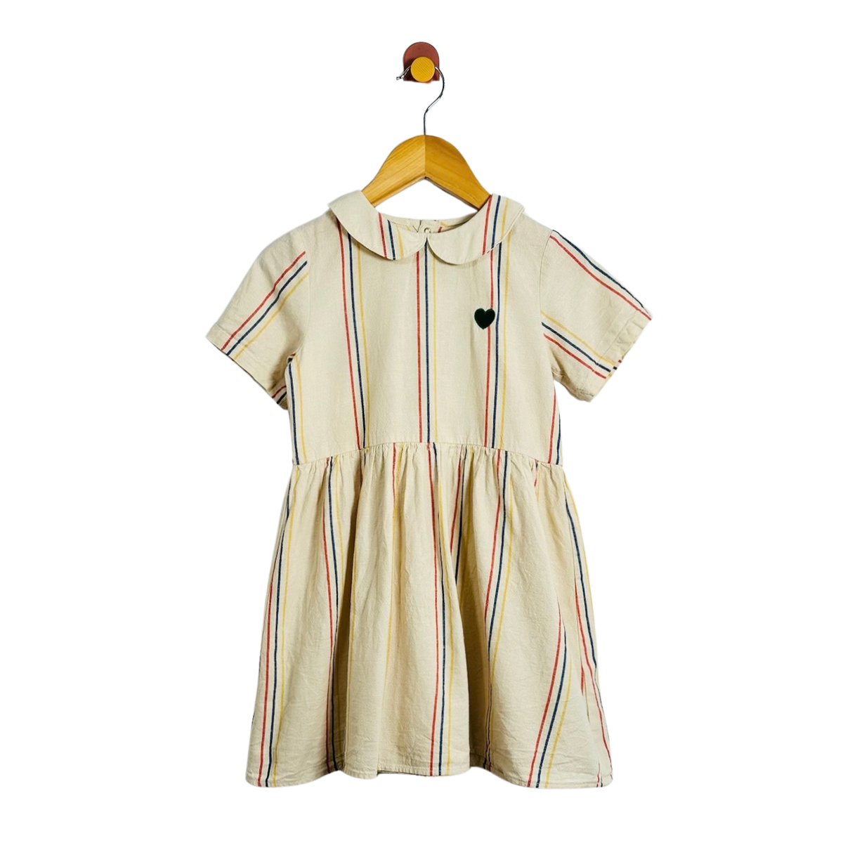 A short-sleeve cream-colored dress with thin vertical multicolor stripes and a small black heart on the chest, hanging on a wooden hanger against a plain white background.