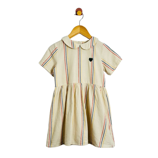 A short-sleeve cream-colored dress with thin vertical multicolor stripes and a small black heart on the chest, hanging on a wooden hanger against a plain white background.