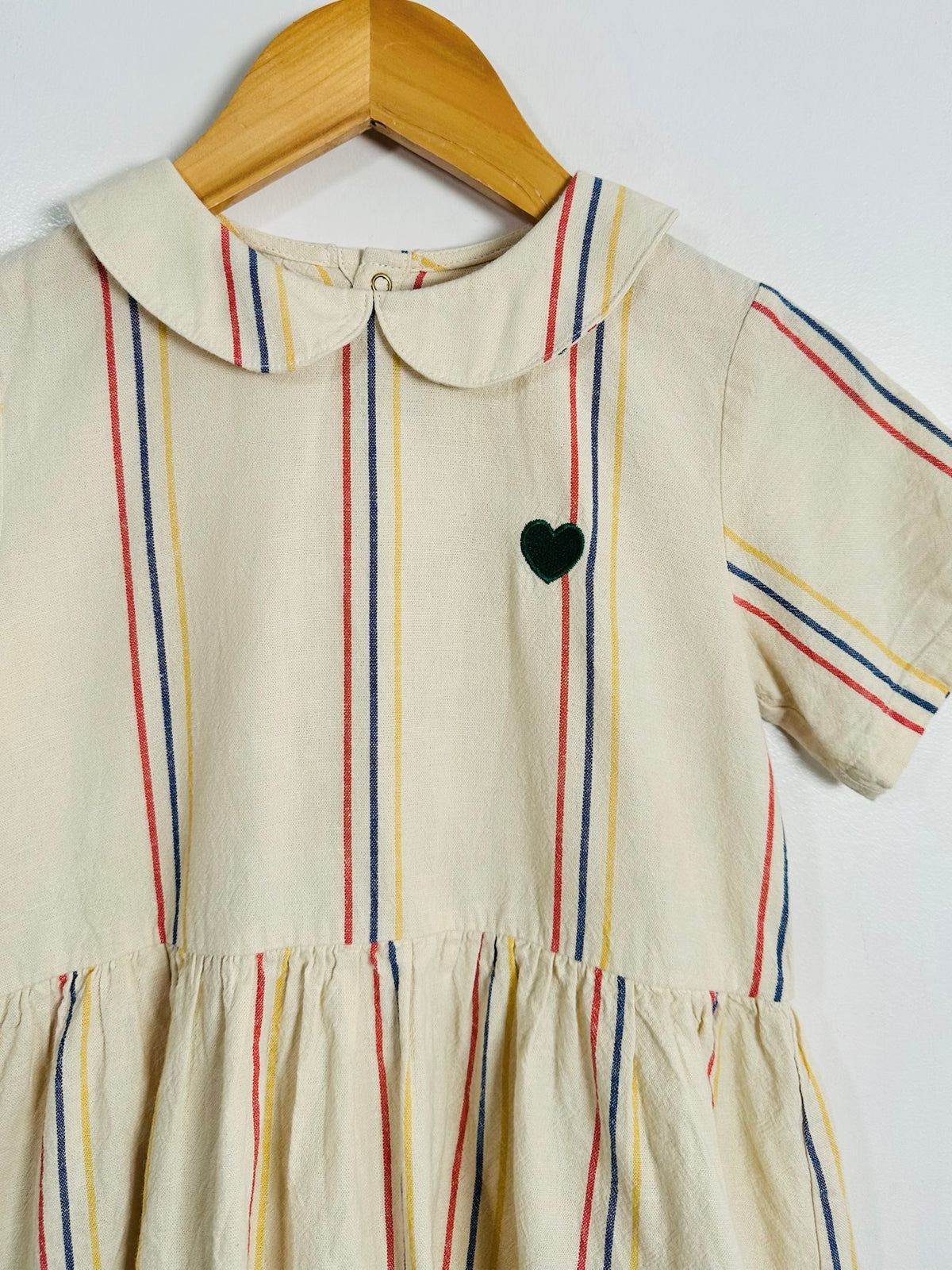 A cream-colored short-sleeve dress with vertical red, blue, and yellow stripes, featuring a Peter Pan collar and a small black heart on the chest, hanging on a wooden hanger against a white wall.