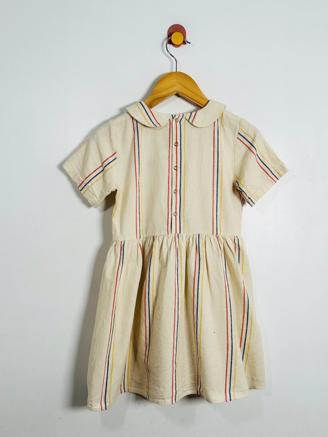 A short-sleeve cream-colored dress with thin vertical red, blue, and yellow stripes, hanging on a wooden hanger against a plain white wall.