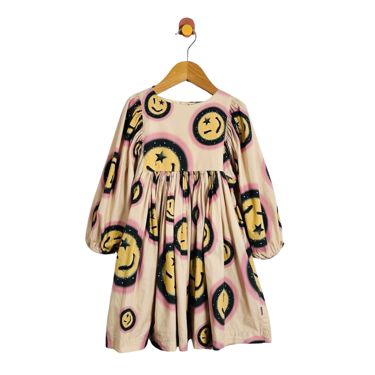 A beige long-sleeve dress with a playful print of yellow smiley faces outlined in black and pink spray-paint splatters, hanging on a wooden hanger against a white background.