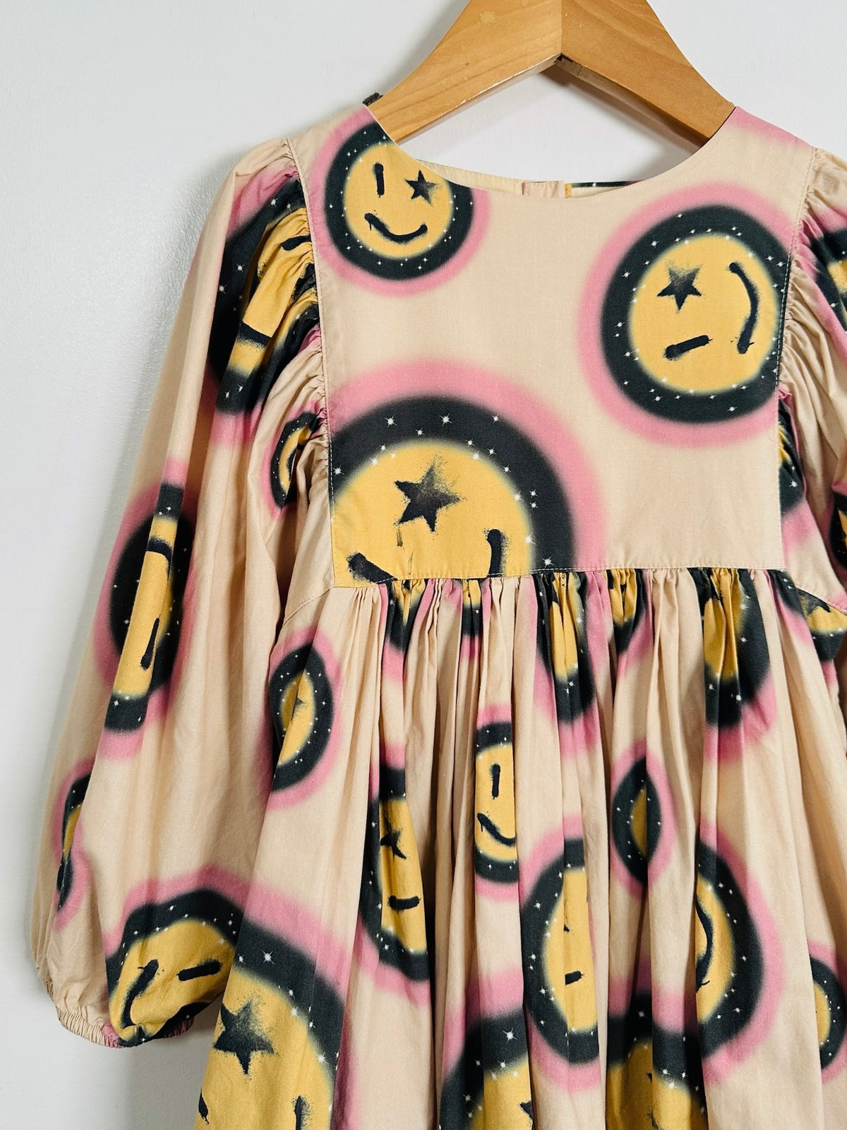 A beige long-sleeve dress with a yellow smiley face print, featuring black star details and pink spray-paint style outlines, hanging on a wooden hanger against a white wall.