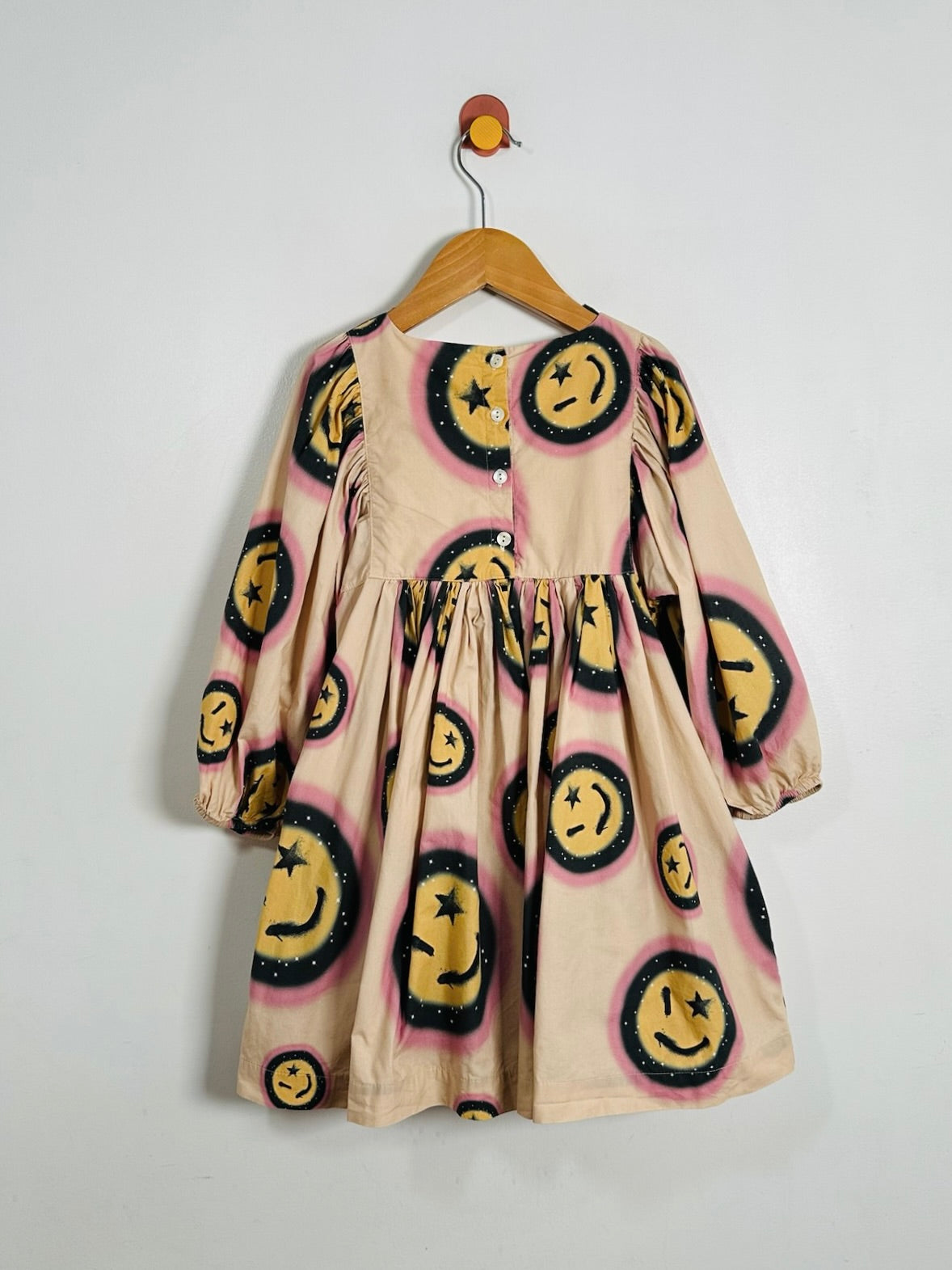 A beige long-sleeve dress with a yellow smiley face pattern, hanging on a wooden hanger against a plain white wall.