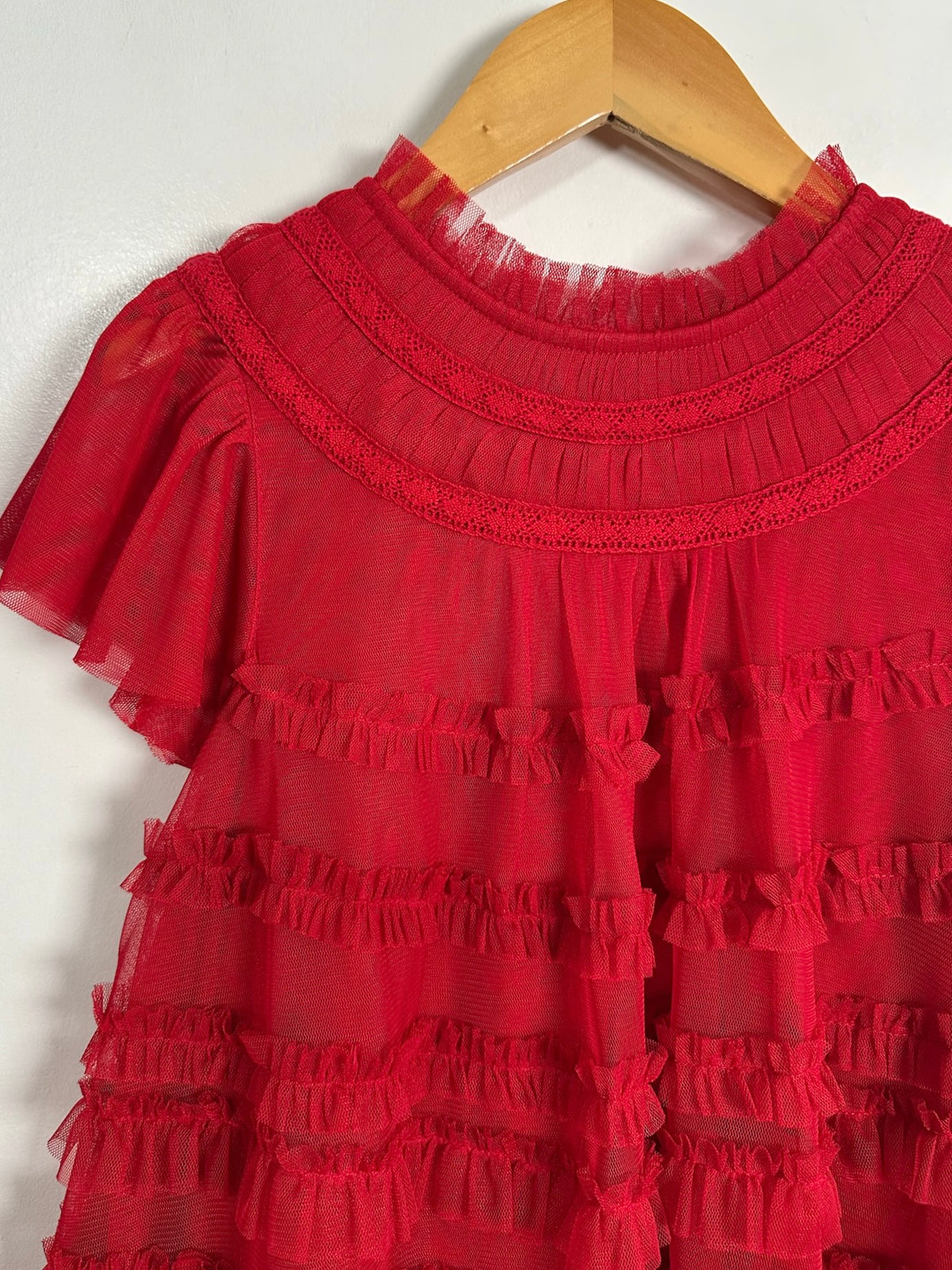 A red tiered tulle dress with ruffles and a high ruffled neckline, hanging on a wooden hanger against a plain light background.