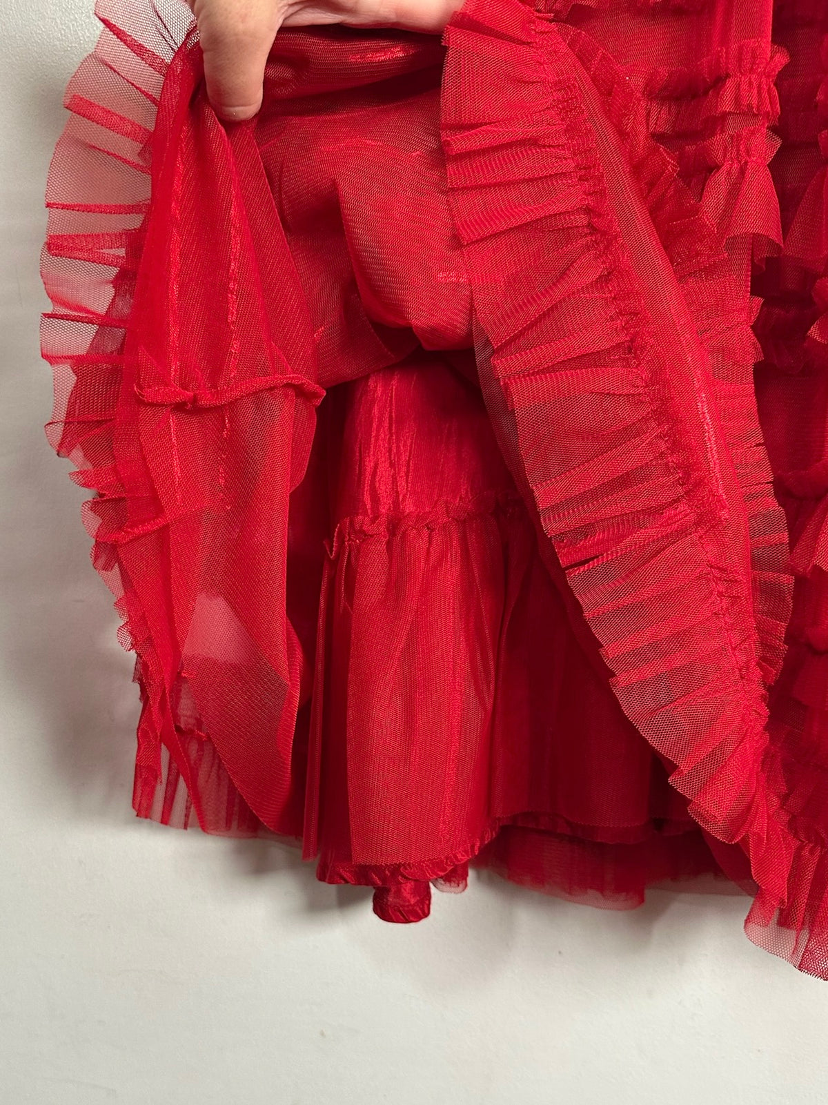 A close-up of a bright red sheer tulle skirt with multiple ruffled tiers, held up by a hand against a plain light background.