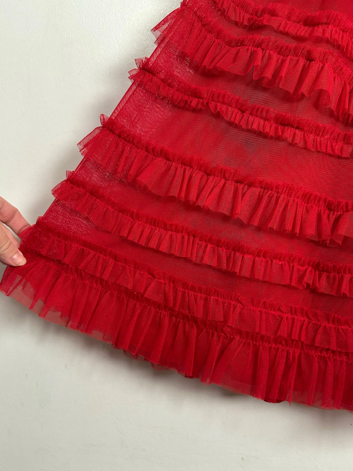 A close-up of a bright red ruffled tulle skirt or dress layer held by a hand against a plain light background.