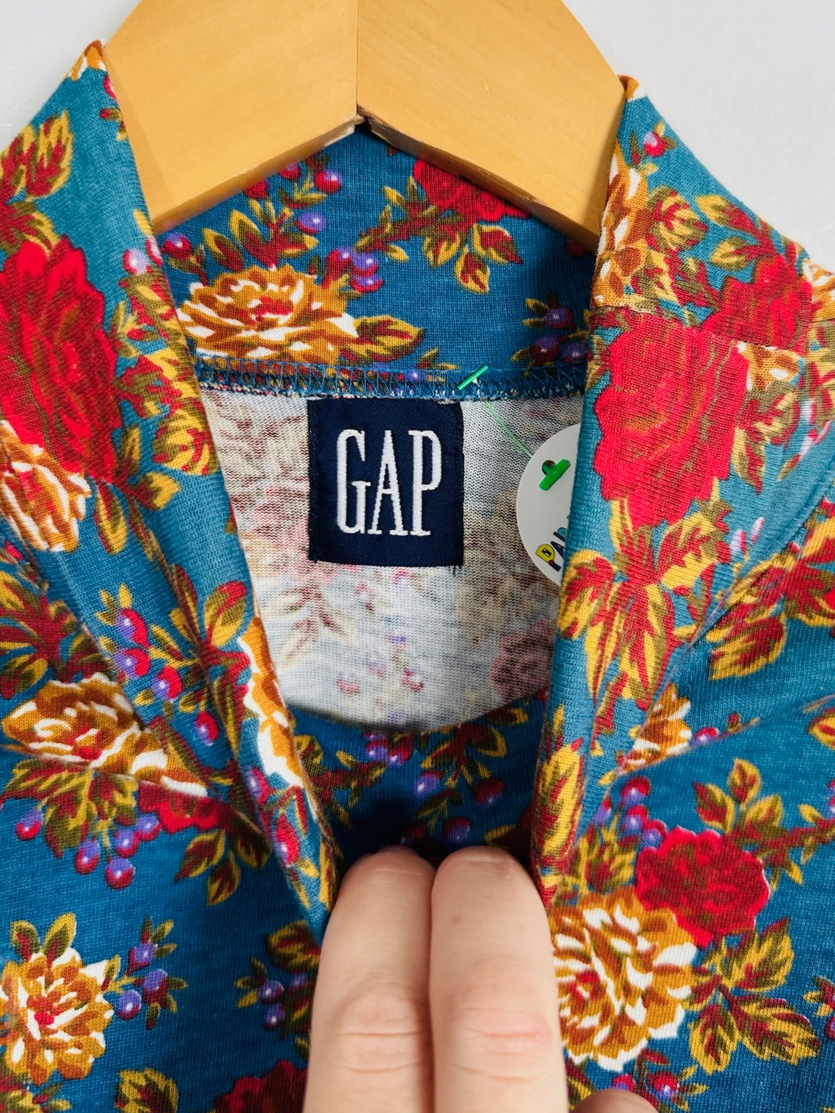 Close-up of a floral-patterned garment on a wooden hanger, showing a visible GAP neck label and a hand holding the collar open.
