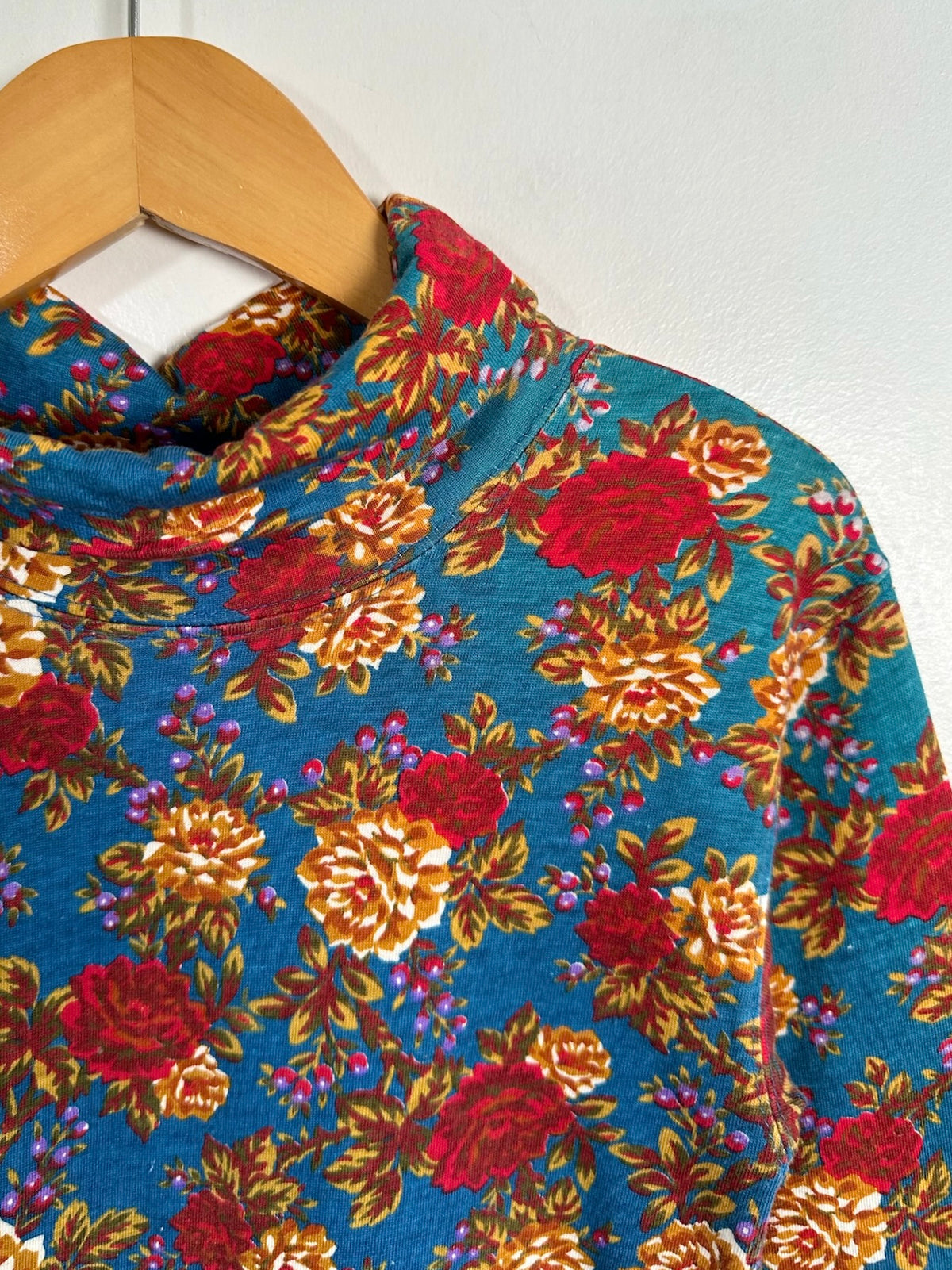 A close-up of a vintage floral turtleneck sweater hanging on a wooden hanger against a plain white wall. The sweater features a vibrant blue base with a retro floral pattern in red, orange, and yellow.