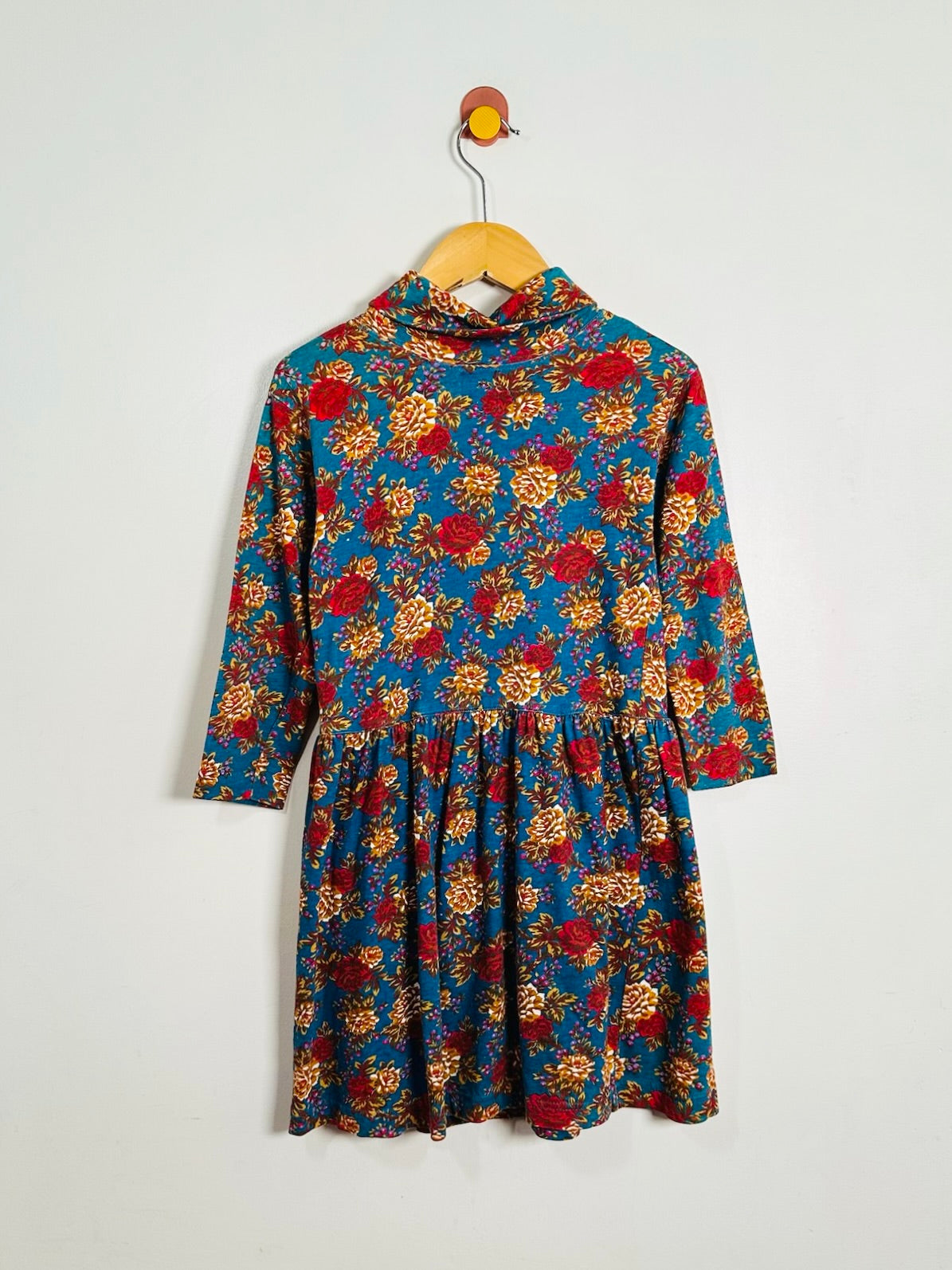 A long-sleeve floral dress with a gathered waist, hanging on a wooden hanger against a plain light gray wall.