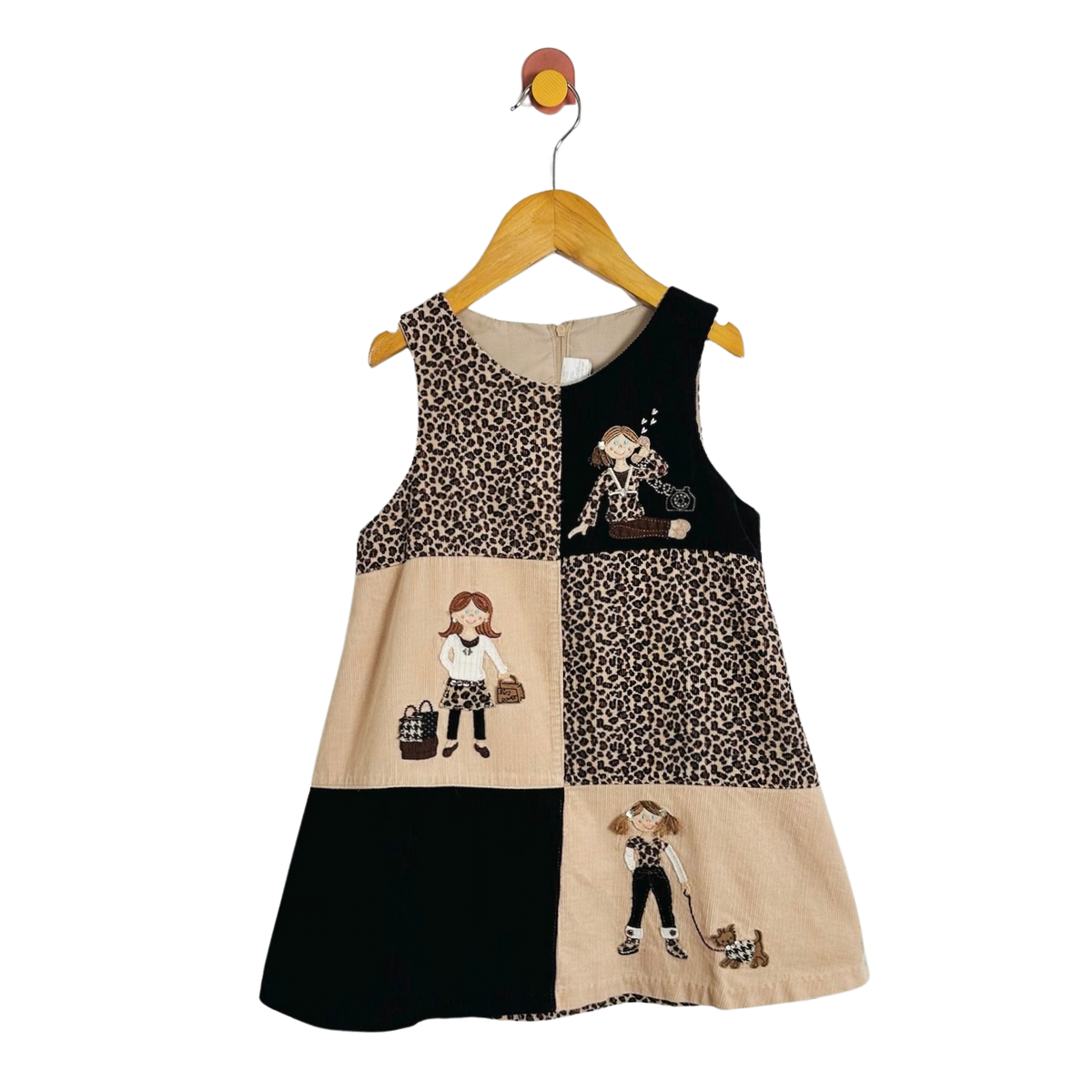 A sleeveless patchwork dress for toddlers, featuring leopard print, beige panels with cartoon illustrations, and solid black sections, hanging on a wooden hanger against a white background.