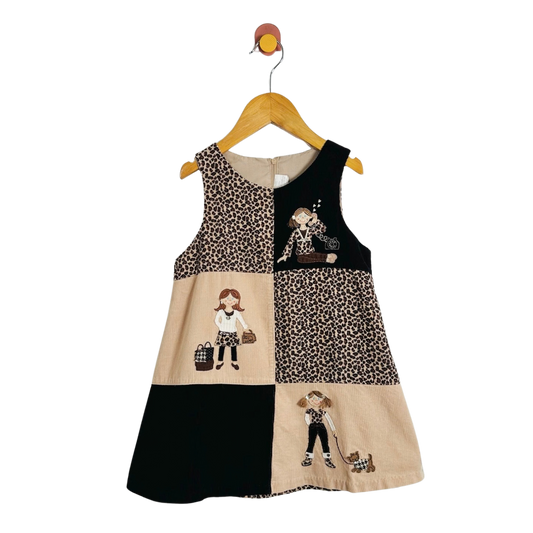 A sleeveless patchwork dress for toddlers, featuring leopard print, beige panels with cartoon illustrations, and solid black sections, hanging on a wooden hanger against a white background.