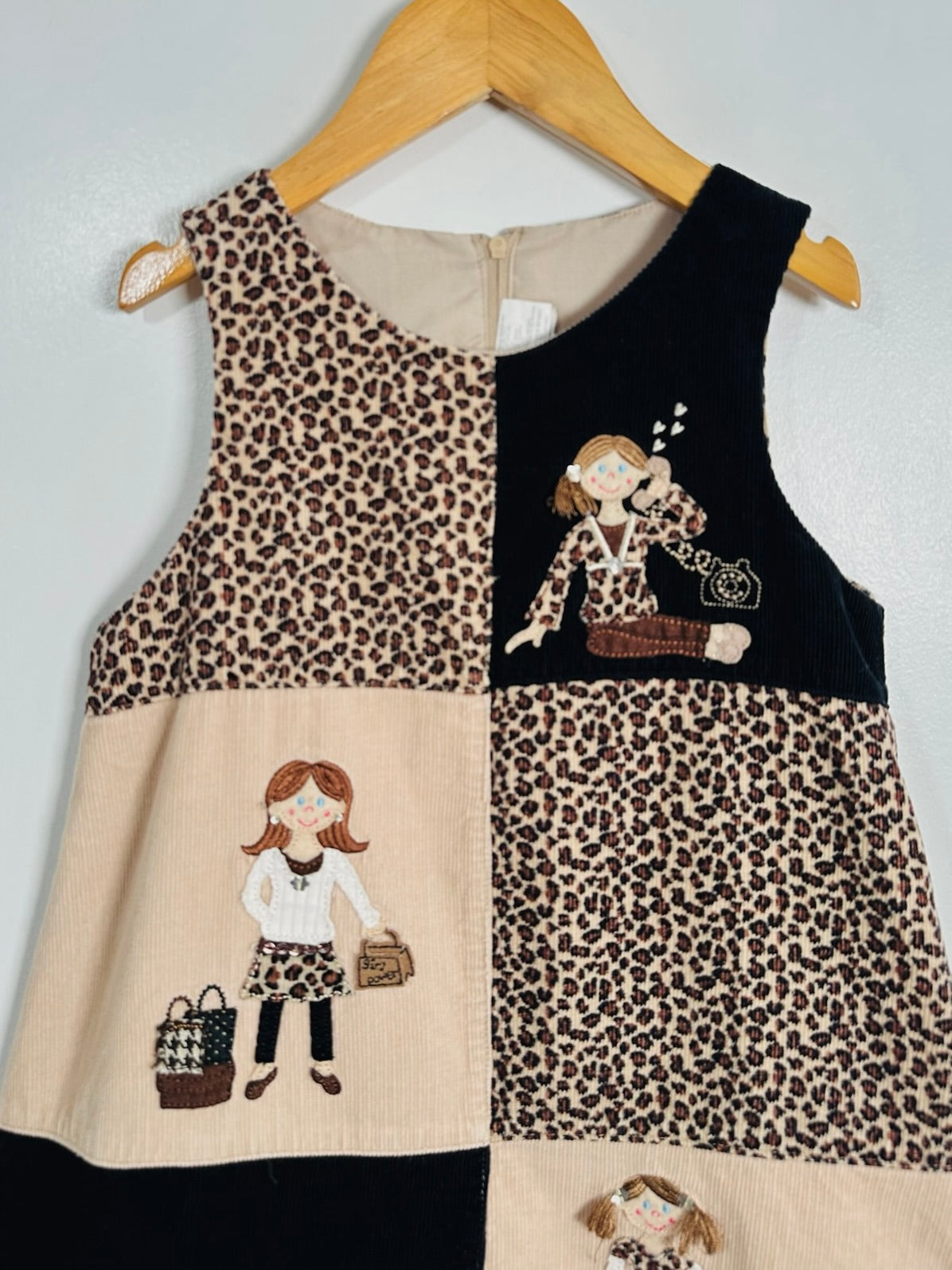 A sleeveless patchwork vest hanging on a wooden hanger against a light gray wall. The vest features leopard print panels, solid black panels, and beige panels with embroidered cartoon girl motifs.