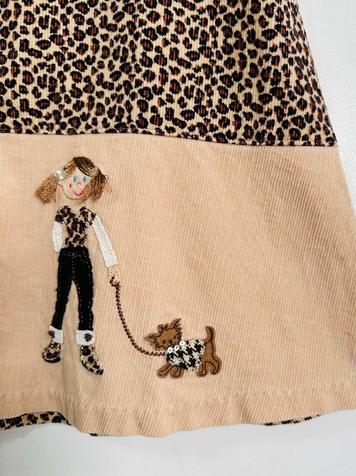 Close-up of a beige corduroy skirt with a leopard-print upper section and an embroidered design of a girl in a leopard-print outfit walking a small dog.