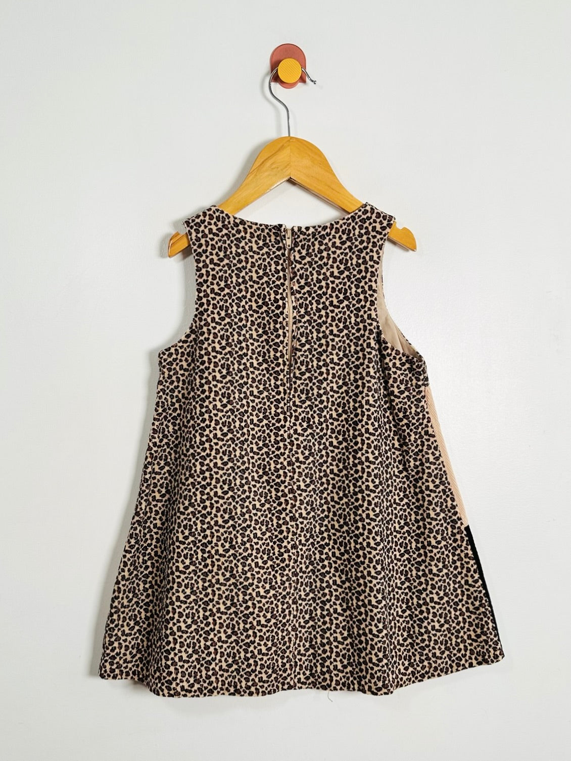 A sleeveless children's dress with a brown and beige leopard print pattern, hanging on a wooden hanger against a plain white wall.
