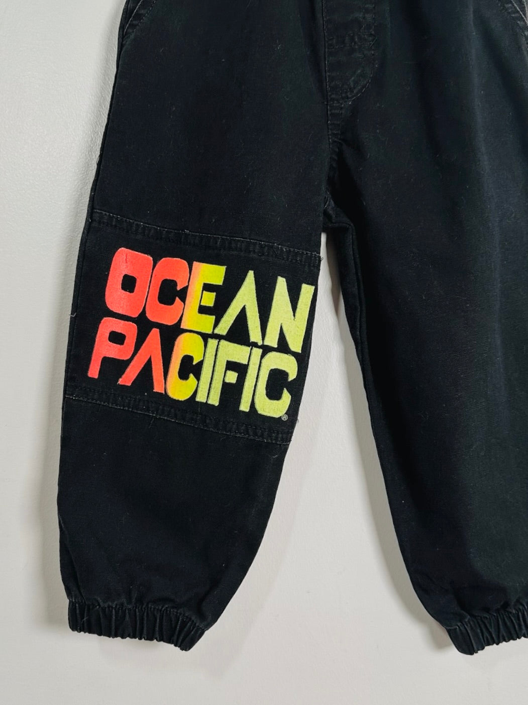Black denim pants featuring a large, colorful 'OCEAN PACIFIC' graphic printed on the left thigh pocket area, shown against a plain light gray background.