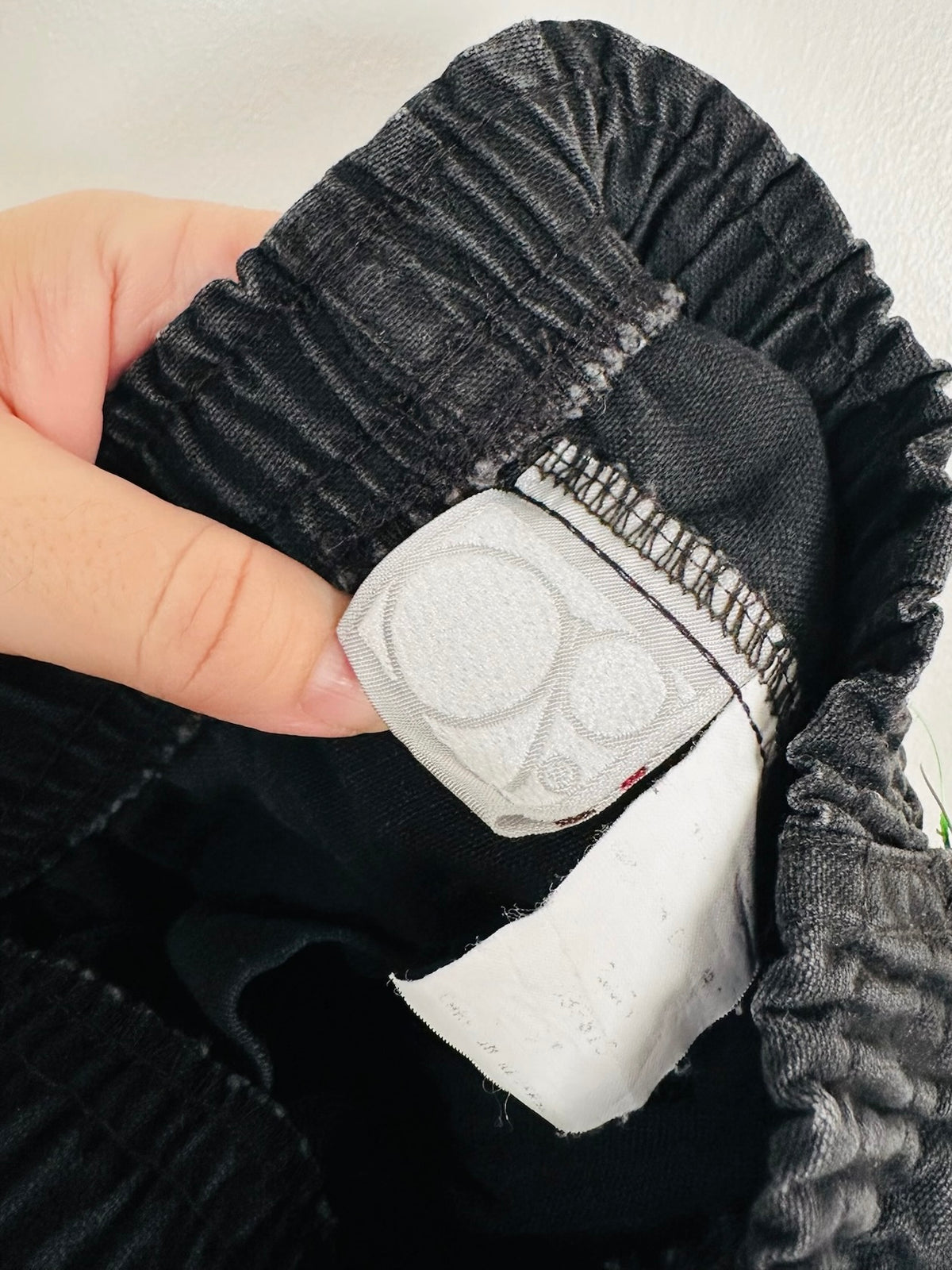A close-up of a person's hand holding a pair of black ribbed shorts, showing a white fabric patch with two circular cutouts sewn onto the inside waistband.