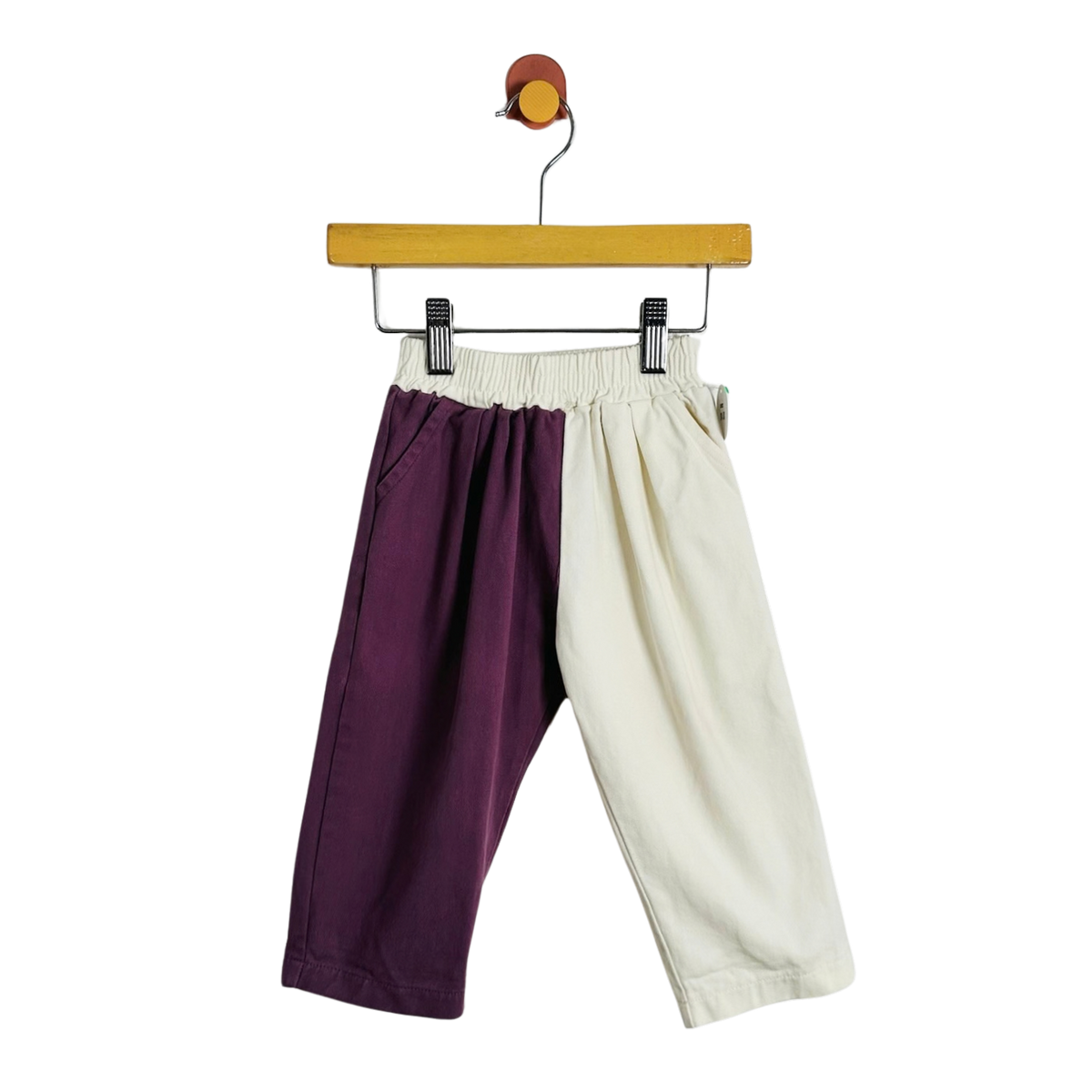 A pair of children's cropped pants hanging from a wooden hanger on a white background. The pants are split down the middle with one leg in deep purple and the other in off-white/cream.
