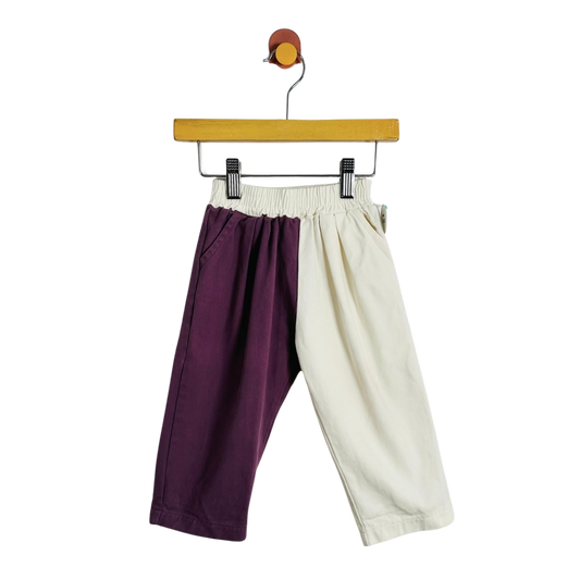 A pair of children's cropped pants hanging from a wooden hanger on a white background. The pants are split down the middle with one leg in deep purple and the other in off-white/cream.