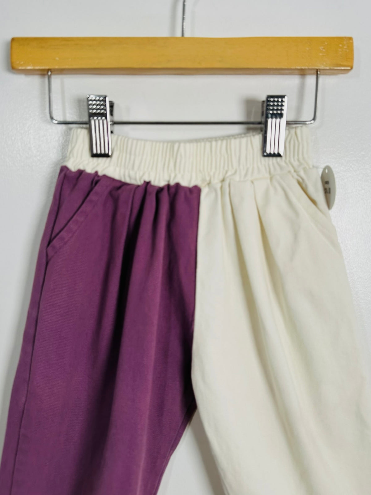 A pair of loose-fitting pants hanging from a wooden hanger with metal clips. The left leg is a deep purple, and the right leg is an off-white/cream color, with an elastic waistband.