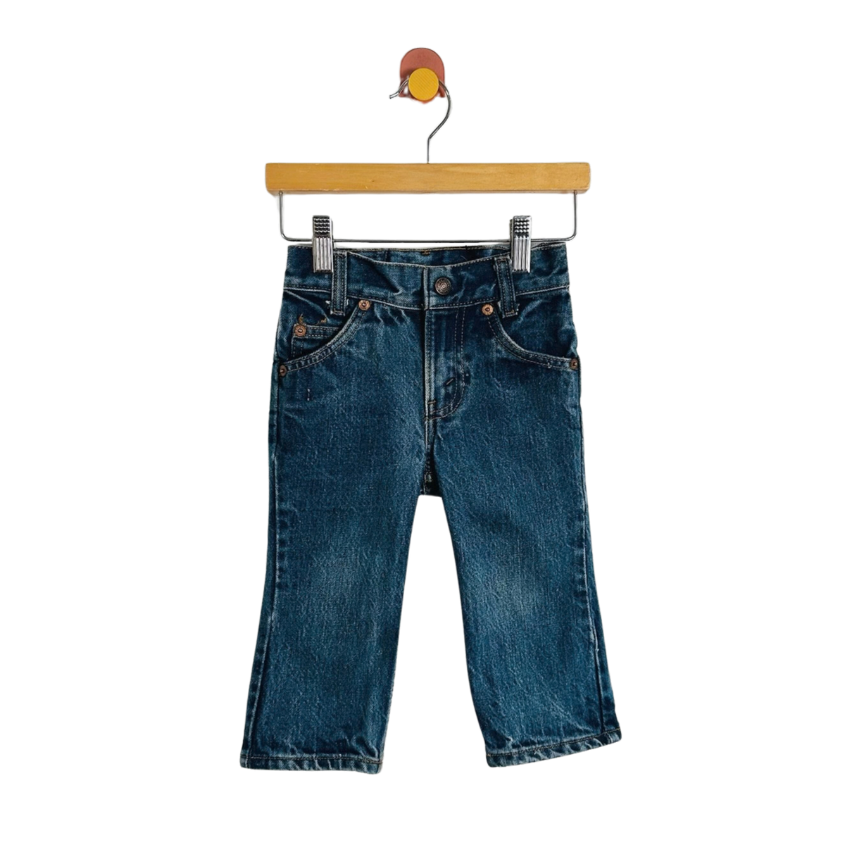 A pair of dark blue denim jeans with a straight, cropped leg, hanging from a wooden hanger with metal clips against a plain white background.