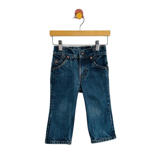 A pair of dark blue denim jeans with a straight, cropped leg, hanging from a wooden hanger with metal clips against a plain white background.