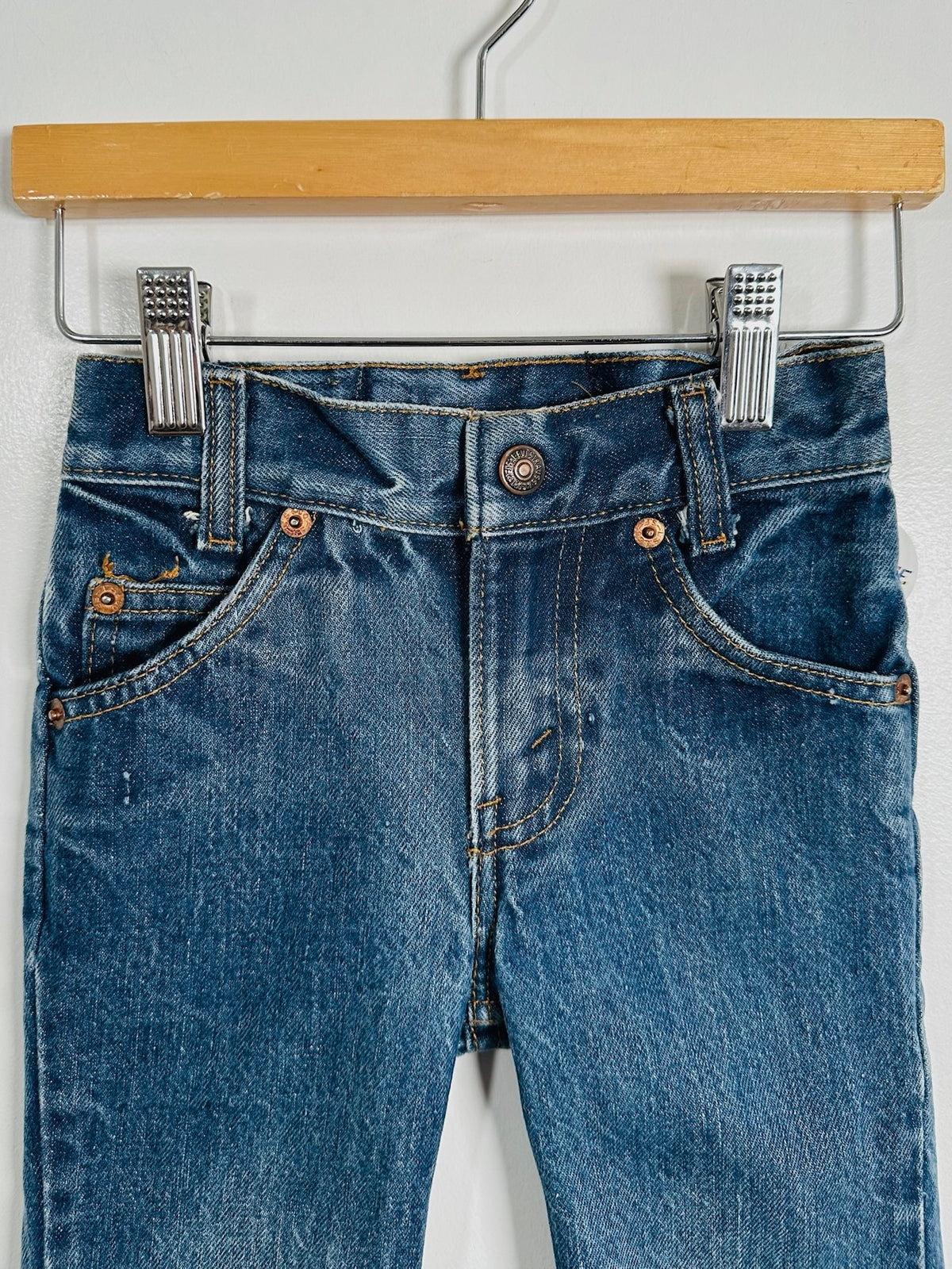 A pair of blue denim jeans hanging from a wooden hanger with metal clips against a plain white wall.