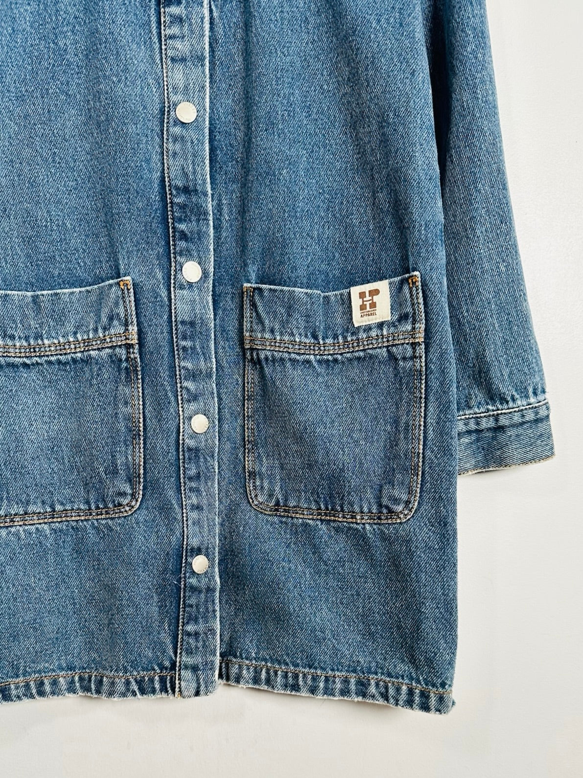 Close-up of a blue denim button-front shirt jacket with two front patch pockets and a small square brand patch on the right pocket, shown against a plain light background.
