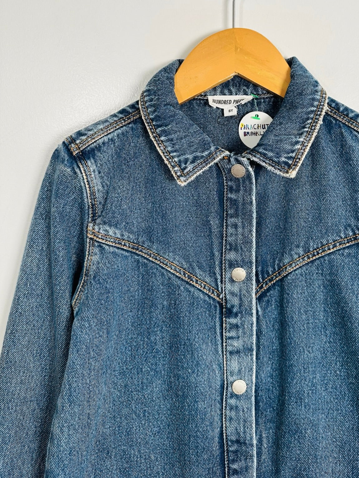 A blue denim western-style shirt jacket hanging on a wooden hanger against a plain light gray wall. The jacket features contrast stitching, a pointed collar, and silver-tone snap buttons down the front.