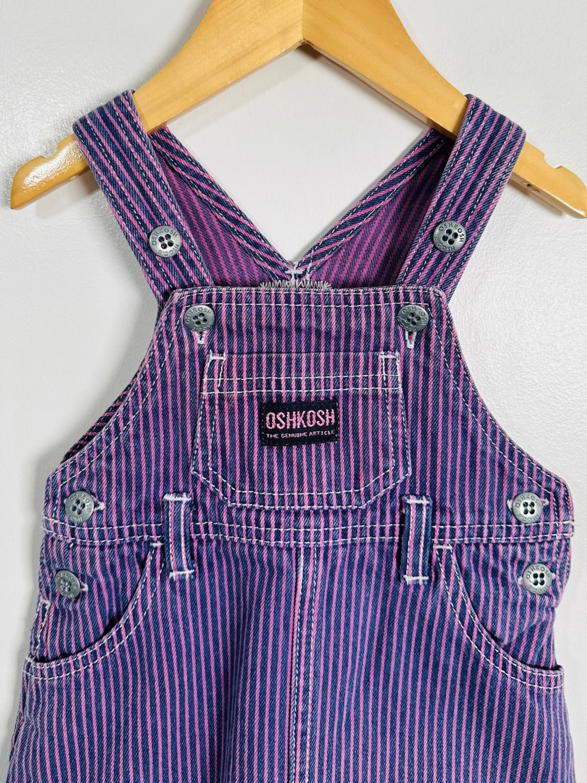 A purple and white striped denim overall garment hanging on a wooden hanger against a white wall. The brand label 'OSHKOSH' is visible on the chest pocket.